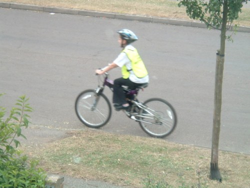 Ein britisches Kind wird im „safe cycling“ unterrichtet. Es trägt fluoreszierende Kleidung sowie einen Helm. Es radelt im Rinnstein einer Wohnstraße mit regem Durchgangsverkehr.