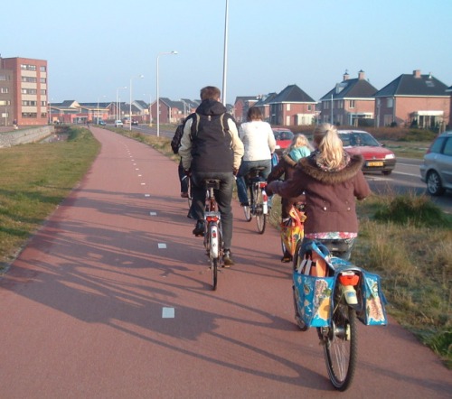 Baulich getrennte Radweg, 4 meter breit, mit ebene, rot-asphaltierte Oberfläsche. Eine Familie fährt Fahrräder auf den Radweg.